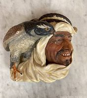 Front and side views of the Bosson head sculpture featuring a man with traditional head covering and a bird on his shoulder.