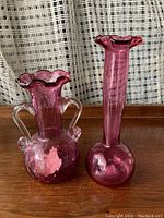 Front view of two cranberry glass vases, one short with handles and one tall with slender neck, on wooden surface with curtain background