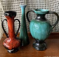 Three large Blue Mountain Pottery vases in red, green, and blue-green glaze, arranged side by side on a wooden surface against a background of white curtains.