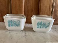 Photo of two matching Pyrex Amish pattern 501 fridge dishes with lids on a marble surface.