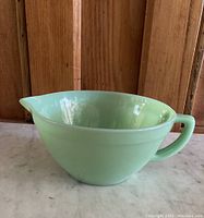 Side angle view showing the light green opaque glass jadite batter bowl with handle and spout, on a wooden background.