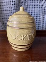 Closed view of vintage barrel-shaped ceramic cookie jar with embossed COOKIES text and ridged bands, cream color glaze.