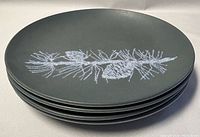 Stack of four dark matte finished plates with white pine cone and pine needle design on top plate.