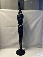 Full view of black painted elongated male metal sculpture with textured surface on white backdrop.