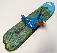 Side angle showing bluebird perched on textured scenic base with green foliage and blue sky