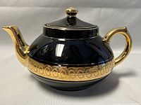 Side angle of teapot showing black glossy body with gold pattern band and gold spout, handle, and lid knob