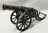 Side view of the metal miniature cannon showing copper barrel and iron carriage with spoked wheels and ornamental supports.