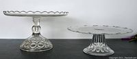 Side view of two clear glass pedestal cake stands with decorative bases and plates, showing scalloped edges and embossed designs.