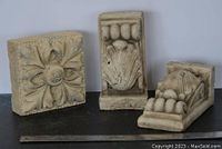 Front view of three cement garden pieces showing one floral tile and two shell motif corbels on a flat surface with a ruler for scale.