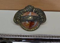 Close-up of one metal drawer pull with ornate scroll and floral design and central marbled Bakelite insert in orange and cream tones.