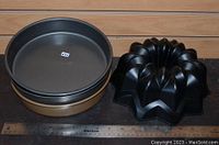 Four baking pans including two dark non-stick 9-inch round pans (one Calphalon marked), one gold-colored round pan, and one black Bundt pan with fluted star design next to a ruler for measurement.
