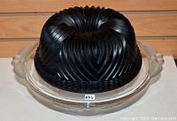 Black bundt pan placed upside down on clear glass cake stand with decorative handles on sides.