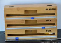 Front view of the wooden dispenser showing compartments labeled Plastic, Wax, and Foil with blue cutter sliders.