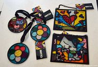 Five colorful Romero Britto style suncatchers displayed on a white surface with attached black ribbons and chains.