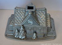Side/top angled view of the silver house-shaped baking pan with detailed roof tiles, windows, door, and trees on the base showing intricate exterior design.
