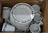 Top-down view of boxed Christmas dinnerware set showing stacks of dinner plates and mugs with holly berry design around rims
