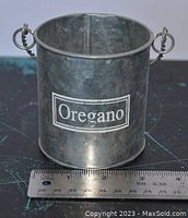 Close-up of a single printed tin pot labeled Oregano with a ruler showing size around 3 inches tall and 3 inches diameter.