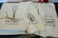 Four printed kitchen towels stacked, showing 'WHIP IT GOOD' with whisk, 'Cheers!' with corkscrew, and 'Eat Drink Be Merry' towel edges.