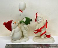 Photo of two Snow Babies figurines, including one snowman with a red hat holding a balloon and a child figurine in a green hat, alongside another child figurine holding a red ribbon and spool, on a white surface with ruler for scale.