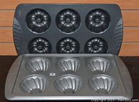 Two Wilton mini Bundt pans, one black and one silver, each with 6 cake cavities, positioned against a wooden surface.