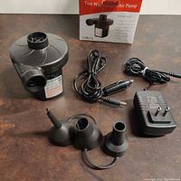 Electric air pump with power cords and three different nozzle attachments displayed on table, original box in background.