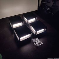 Four black square solar-powered post lights lit up on a dark surface, showing white light from translucent panels. A small plastic bag with mounting hardware is placed near the lights.