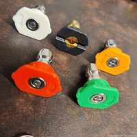 Five power washer replacement tips arranged together on a surface. Color-coded nozzles: red, green, orange, white, and black with metal connectors.