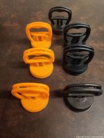 Top and angled view of four black suction cups and three orange suction cups arranged on a dark surface, showing their handles and bases.