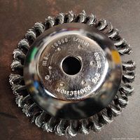 Photo showing top view of the stainless steel brush clearing disc with twisted wire bristles and a central mounting hole with protective cover and embossed text.
