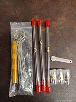 Set of tubes containing airbrush needles, labeled by size, with red caps, alongside a gold handle, wire cleaning brushes in a plastic bag, a small wrench, and small plastic containers.
