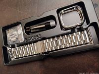 Top-down view of the silver stainless steel Apple iWatch bracelet inside black plastic storage tray. Includes pin pusher tool, extra links, and matching frame.