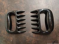 Two black plastic meat shredding claws resembling bear paws shown side by side on a dark surface.