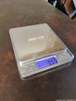 Compact digital scale powered on showing zero reading on blue backlit LCD display, silver surface, three buttons on front.