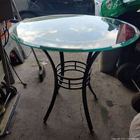 Side view of round glass top table with black metal legs, showing the curved leg design and circular grid structure connecting legs near the base.