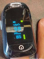 Front view of the black fingertip pulse oximeter showing digital screen with SpO2 reading of 98%, pulse rate 85 BPM, battery level, and heart icon.