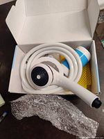 Open box showing a white handheld showerhead and white flexible hose coiled together inside the box, near a piece of bubble wrap.