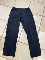 Full view of one pair of navy tactical pants laid flat on tile floor showing front side, including multiple cargo pockets and D-ring on side.