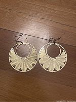 Pair of large round gold-toned metal earrings on wood floor showing detailed petal cutout design