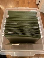 Overhead view inside clear plastic filing box showing multiple green hanging file folders with white labels.