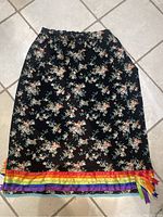 Full view of the skirt laid flat, showing black floral fabric with rainbow ribbon stripes at bottom edge.