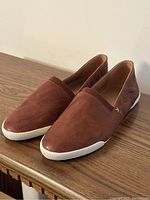 Pair of brown leather slip-on shoes with white rubber soles positioned on a wooden surface, showing a side and front view.