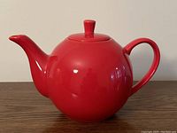 Front side angle showing red ceramic teapot with lid, curved spout and handle lying on wood surface.
