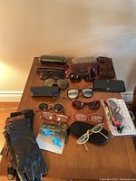 Full view of gloves, sunglasses, reading glasses, sun guards, and cases arranged on a wooden table.