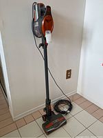 Full view showing Rocket Shark vacuum mounted on the wall upright, showing stick design, dust container, power cord coiled on floor