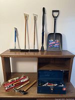 Blue handled floor scraper, two wood handled scrapers with brooms and brush, large plastic shovel, and blue metal toolbox on wooden table.