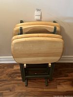 Four wooden TV dinner tables stacked and folded against a wall showing carry handles on tops.