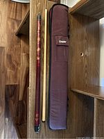 Top down photo of two-piece vintage pool cue and maroon padded carrying case with Cooper logo placed on hardwood floor.