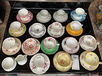 Top-down view of 15 assorted vintage porcelain tea cups and matching saucers arranged on a tray showing variety of floral patterns and colors, including pink, yellow, green, orange, and blue designs.