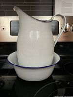 Front view of white enamel jug placed inside the bowl showing the smooth curved handle, spout, and blue rim detail with a clean backdrop of a stove.