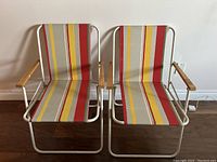 Front view of two vintage folding beach chairs with striped fabric and wood armrests against a white wall and hardwood floor.